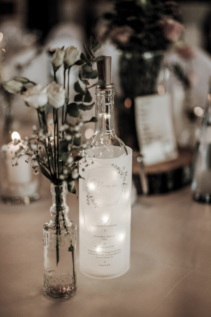 Toller Schmuck für eine Hochzeit bei uns in Günz im Feststadl, zu sehen sind Blumen und eine beleuchtete Weinflasche.