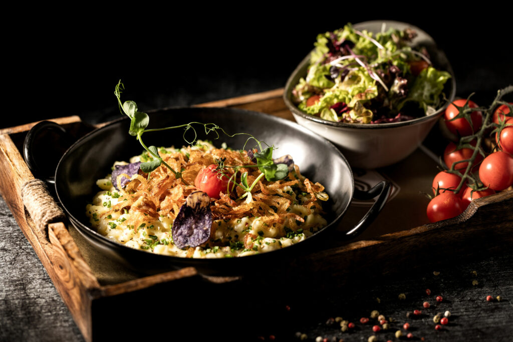 Kässpatzen mit Salat, toll angerichtet und fotografiert auf einem eleganten Holzbrett auf dunklem Hintergrund.