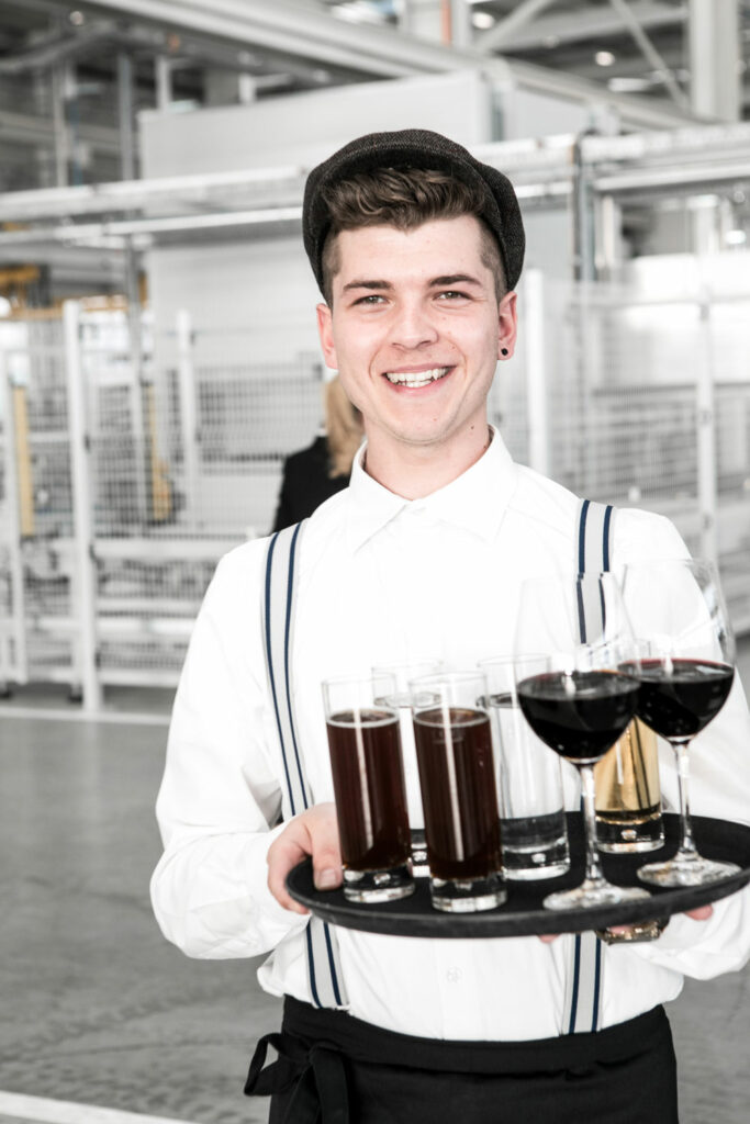 Ein glücklicher junger Kellner beim Catering der Firma Liebherr serviert Getränke und trägt ein schickes Outfit mit Hosenträgern und Schiebermütze.