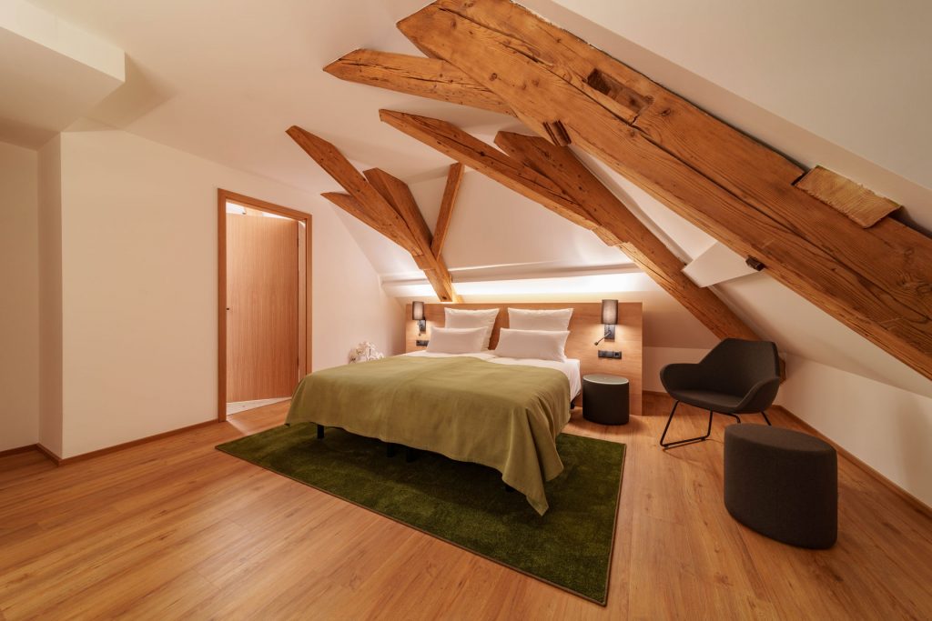 Helles Dachgeschoss-Hotelzimmer mit freigelegten Holzbalken im Brauereigasthof Laupheimer in Günz im Allgäu, nahe Flughafen Memmingen – modernes Design, großzügiger Raum und hoher Komfort.